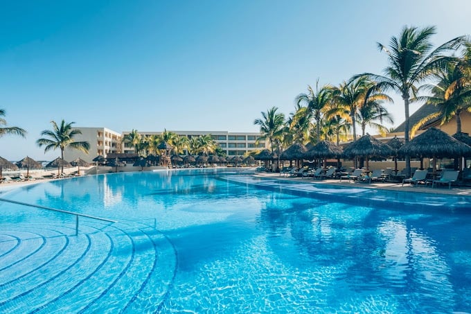 Large luxury resort pool with crystal blue water, palm trees, lounge chairs with umbrellas, and hotel buildings under clear sky