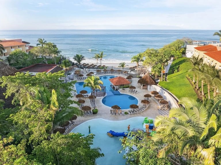 Aerial view of a tropical resort with curved pools, palm trees, beachfront, and ocean in the background