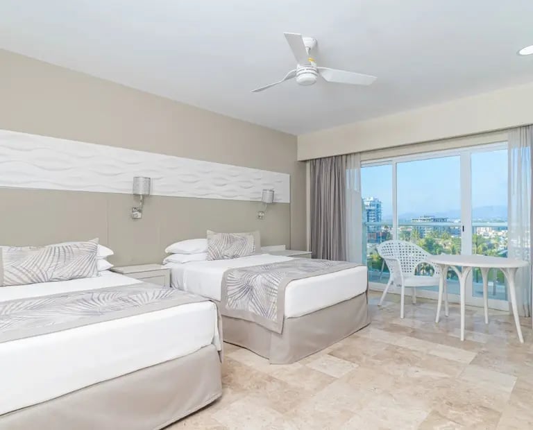 Modern hotel bedroom with two queen beds, beige walls, ceiling fan, large windows with city views, and seating area