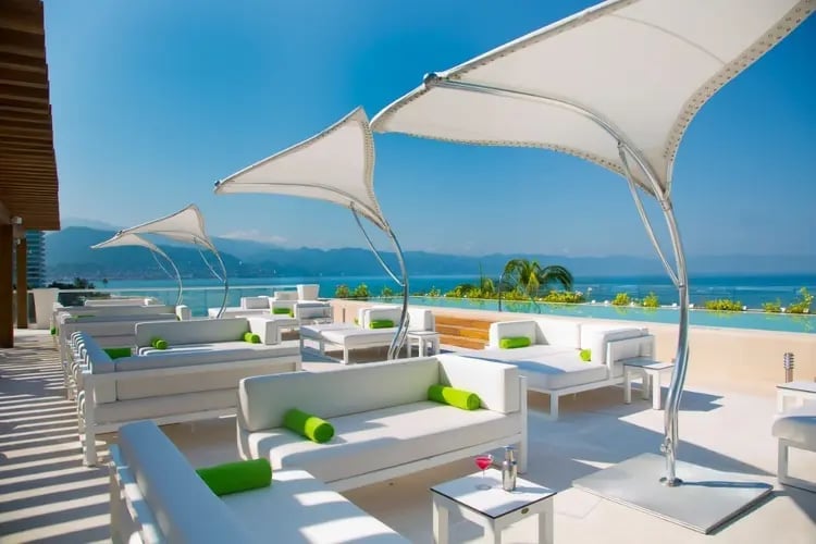 Modern rooftop lounge with white shade sails, contemporary seating, and panoramic ocean and mountain views on a sunny day