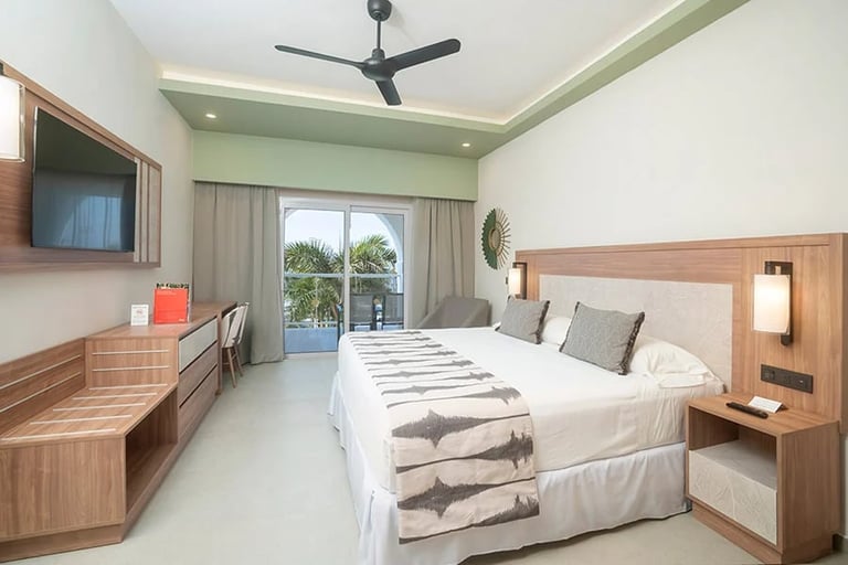 Modern bedroom with queen bed, wooden furniture, wall-mounted TV, ceiling fan, and balcony access with ocean views