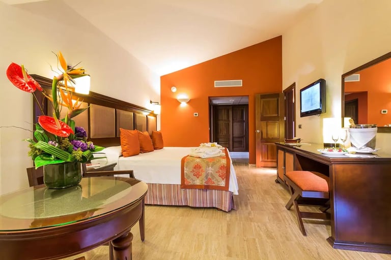 Luxury hotel suite with orange accent wall, wood furnishings, bed with orange bedspread, round table with flowers, and sitting area
