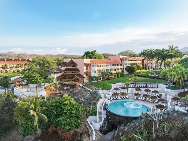 Aerial view of a resort with circular pool, loungers, palm trees, and multi-story buildings surrounded by landscaped grounds