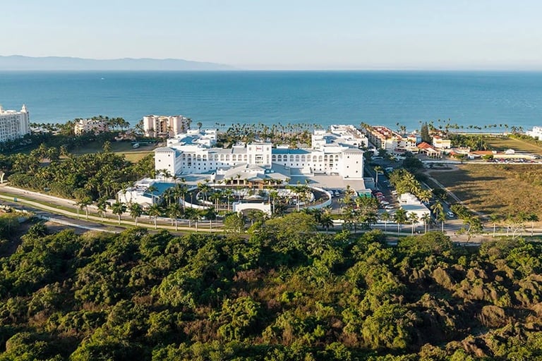 Aerial view of a coastal resort with white buildings, palm trees, and Mediterranean Sea, surrounded by green vegetation