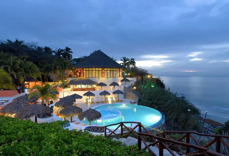 Luxury coastal resort at dusk with illuminated buildings, curved swimming pool, palm trees, and ocean view at twilight