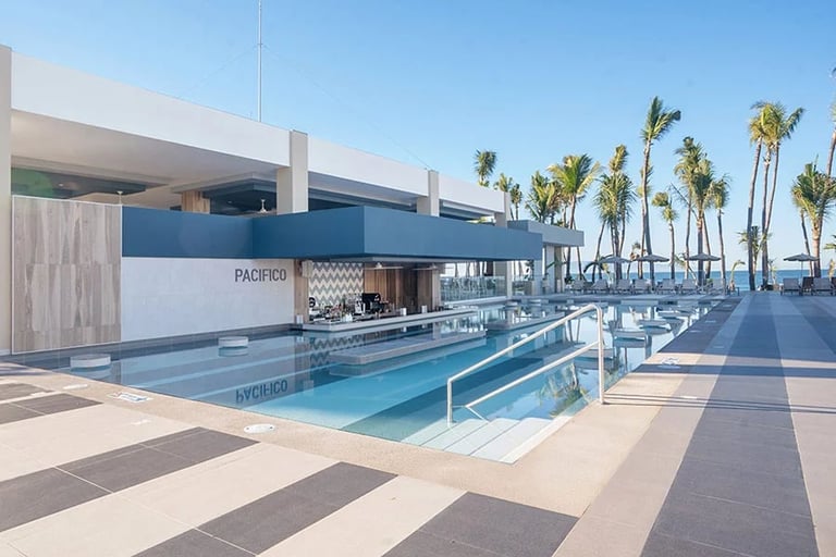 Modern resort pool deck with blue water, lounge chairs, palm trees, and a contemporary poolside building under clear sky