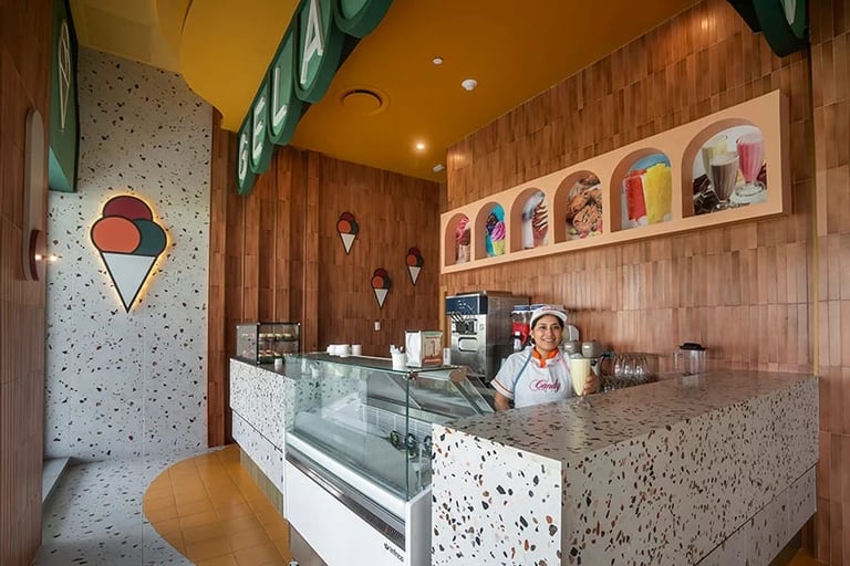 Interior of modern ice cream shop with terrazzo counter, colorful wall shelves, wooden accent wall, and staff member at register