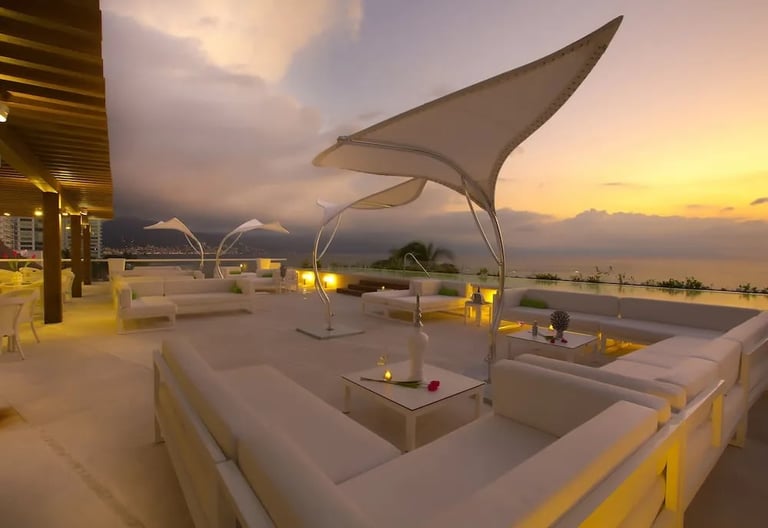 Modern rooftop terrace at sunset with white lounge seating, geometric sail canopy, and water features overlooking the ocean