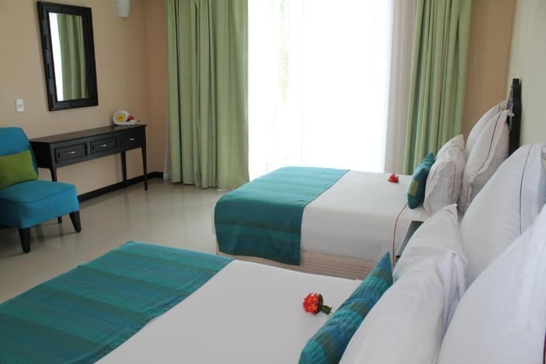Modern hotel room with two beds featuring white and teal bedding, green curtains, desk, chair, and mirror