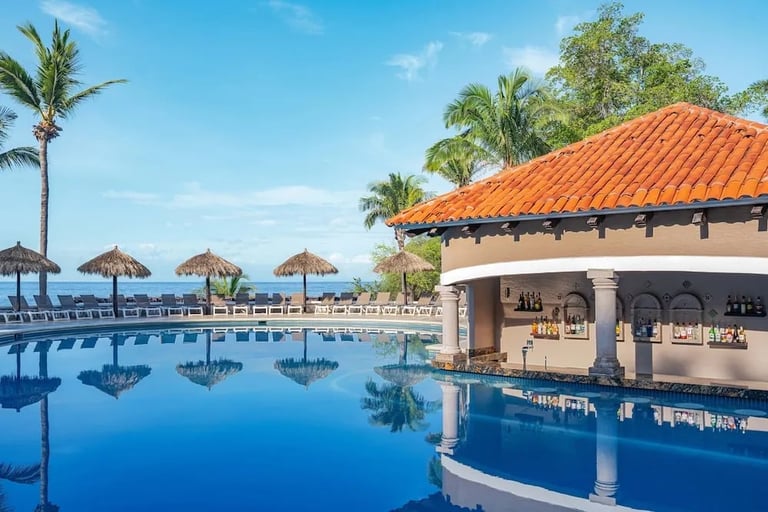 Luxury resort pool with thatched umbrellas, palm trees, and a beachfront pavilion with terracotta roof under clear blue sky