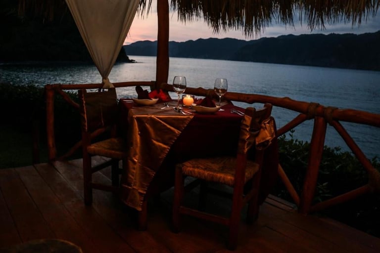 Candlelit outdoor dining table on a wooden terrace overlooking a calm lake at dusk, with thatched roof and tropical setting