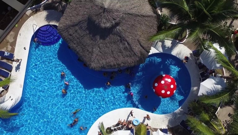 Aerial view of resort pool with turquoise water, thatched umbrellas, palm trees, and red inflatable ball