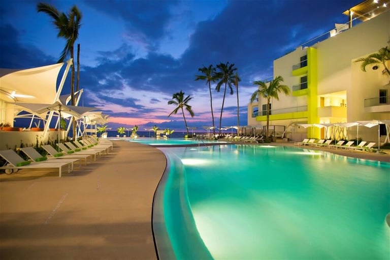 Luxury resort pool at twilight with turquoise water, palm trees, illuminated yellow building, and lounge chairs at dusk